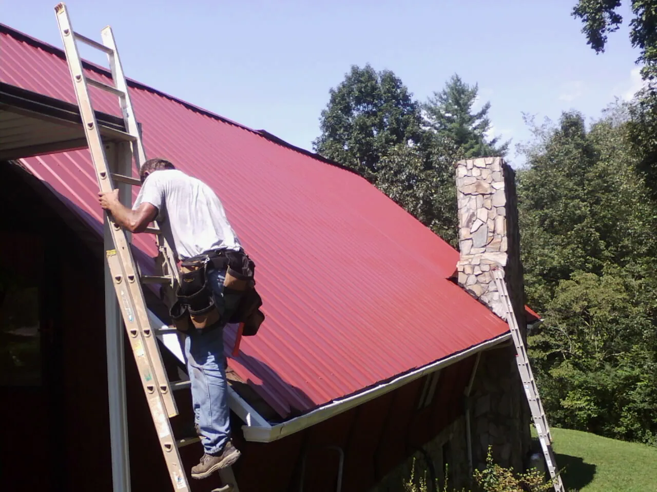 Roofer on a ladder beside a red metal roof and stone chimney — Soffit & Fascia Repair by Roofing Brothers in Otho